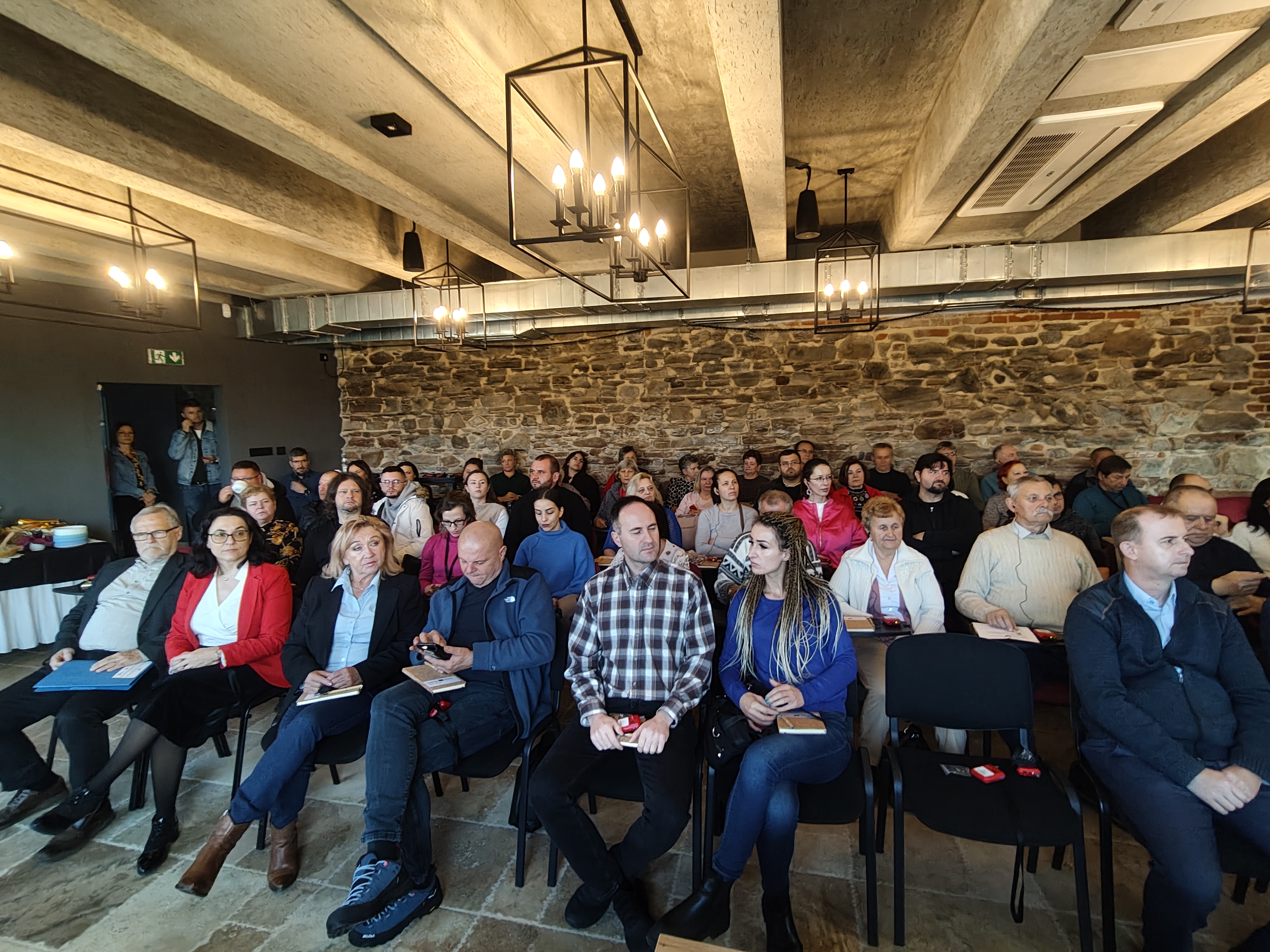 Grupa ludzi siedzi na krzesłach w sali z kamienną ścianą i oświetleniem w stylu loft. Uczestnicy konferencji lub spotkania.